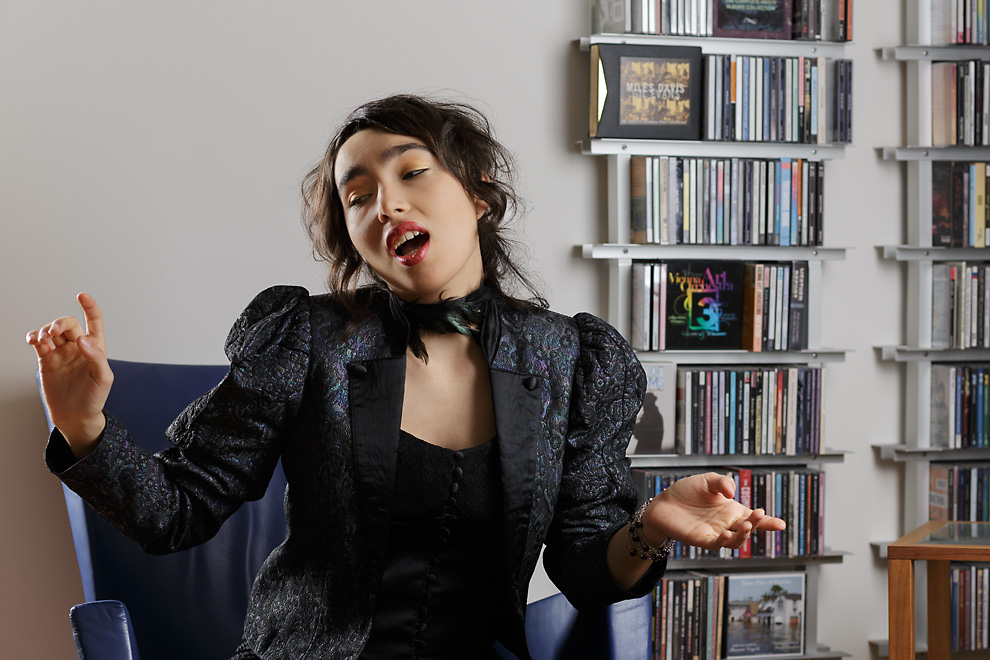 Portrait of a young woman wearing a jacket with puff shoulders, a black top and a feather necklace. She wears wine-red lipstick and golden eyeshadow. She holds her head tilted and looks outside of the picture frame. In the background is a rack with CDs.