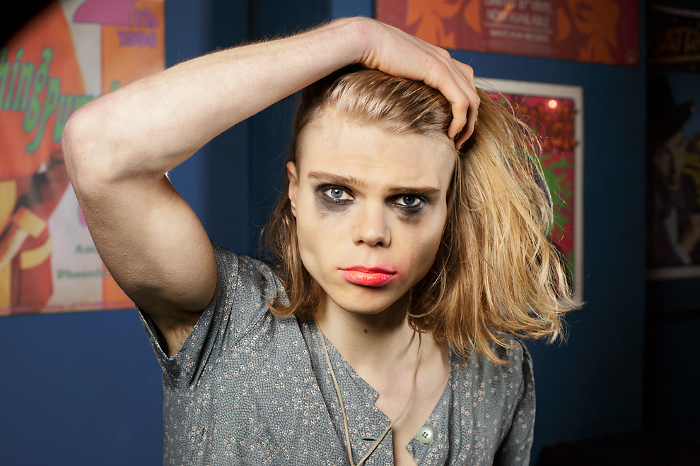 Portrait of a young man, who grabs his longer blond hair on top of his head. He wears a blue-grey-ish dress with a print of small flowers. Both his red lipstick as his black eye shadow are smeared. In the background are gig posters on a blue wall.