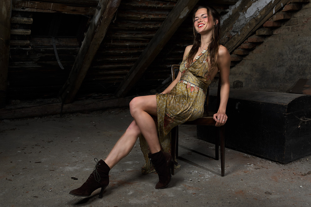 Portrait of a young woman sitting on a chair in an attic. She sits on the edge of the chair and has her right leg strechted out while she leans slightly backwards. She smiles which shows her white teeth framed from red lips. On the dusty floor in the back is an old trunk. She wears a green-gold shiny dress.