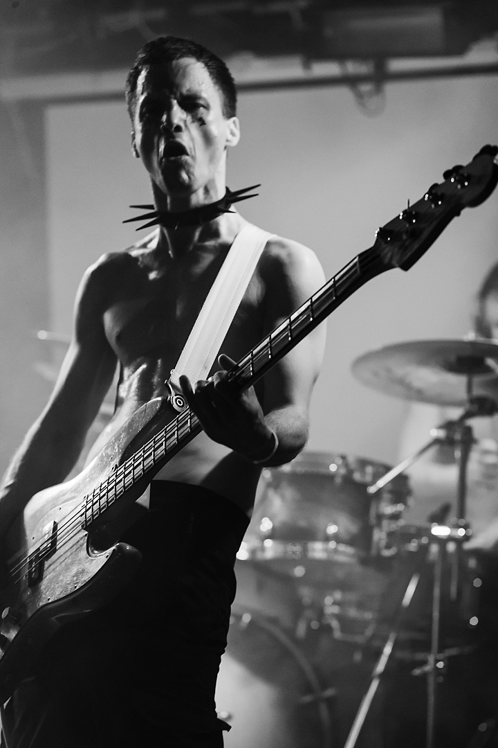 Black and white picture of the bass player of Chants de Rats. Concert photography of a bassist wearing a spiked collar. The drummer is slightly visible in the background.