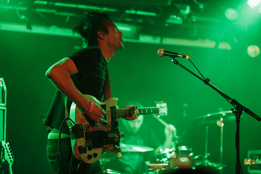 Concert photography of a live performance of Melt Downer. The singer-guitarist is head banging while playing a double-neck guitar. The guitar is customised. The lower neck is sawn off.