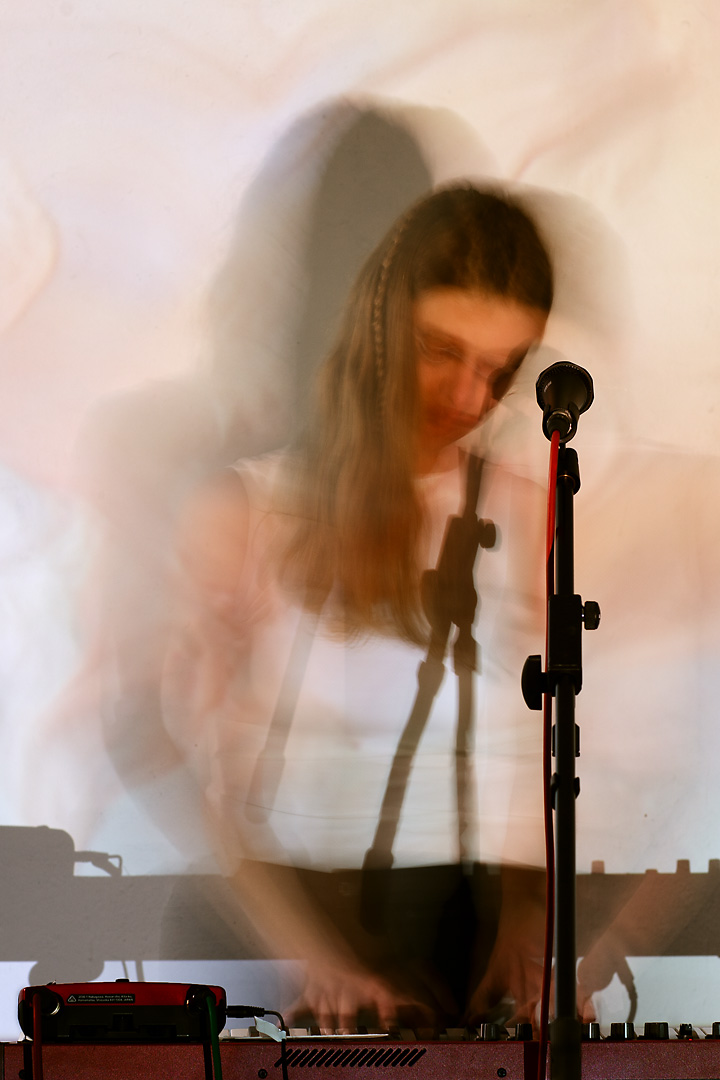 Long exposure picture of Mme Psychosis at a live concert. While static elements such as microphone and synthesizer are sharp, the musician and her shadow are blurry and merge with the background visuals. 