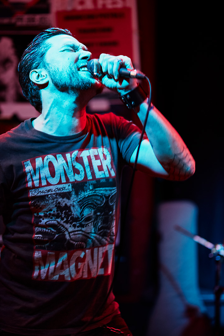 Concert photography of the singer of Out of Nothing. The male singer wears a black Monster Magnet T-shirt and screams in a microphone. His face is lit up by blue light, whereas the background light and strip light is red.