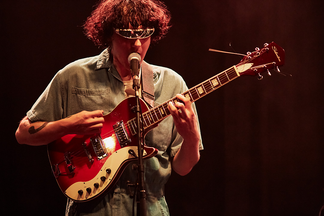 Concert photography of the singer-guitarist of Sux Sux Sux. The guitarist wears blue overalls and space sunnglasses