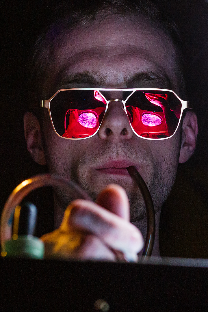 Concert photography of Weird Visuals. A man blows air on a colourful liquid, which is projected in his sunglasses.