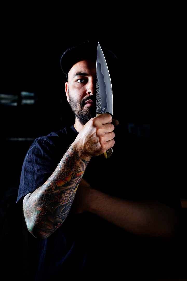 Portrait photography of a Christoph Stieger of Kanji Knives. His black t-shirt and baseball cap blend in with the dark background. Only his right side is lit by a spot. In front of the left side of his face he holds a knife that he crafted by hand. The edge of the knife contrasts with the dark background.