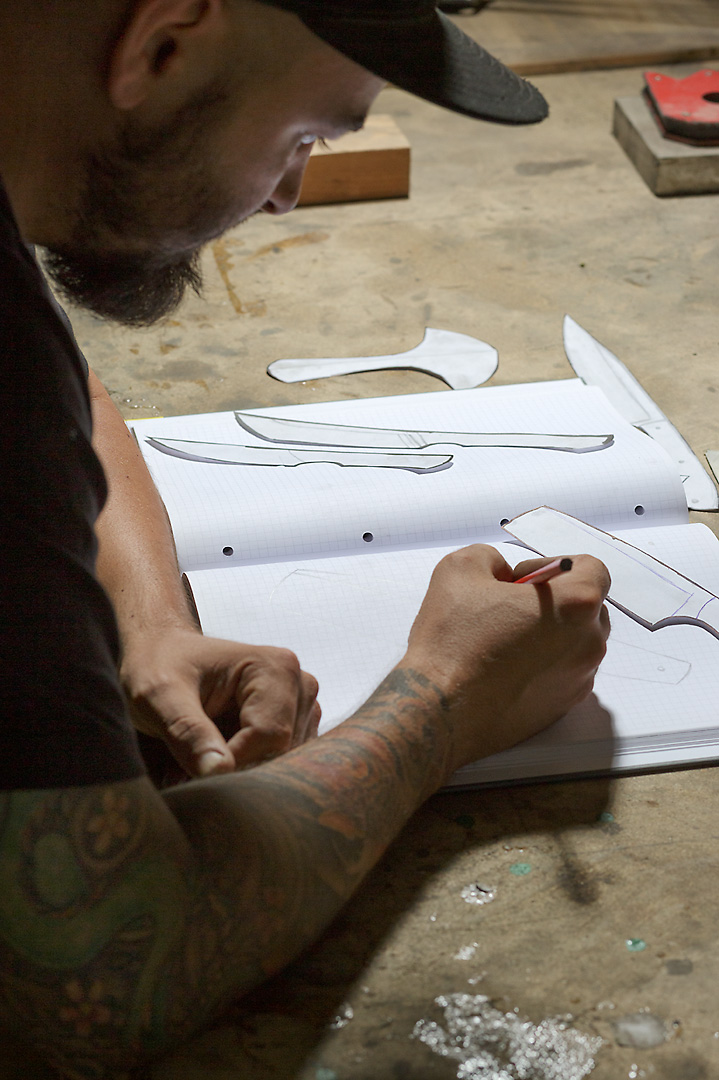 Portrait photography of Christoph Stieger while he designes paper cut-outs of knives. The head and right arm are cropped by the top and left frame. In the center of the picture is a sketch book and paper cut-outs of knives.