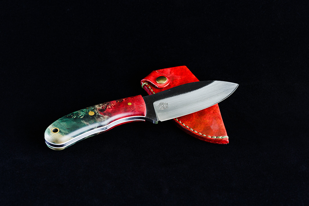 Product photography of a small knife with the Kanji Knives logo ingraved on a black background. The handle is green and red. The knife rests on a red leather sheath.