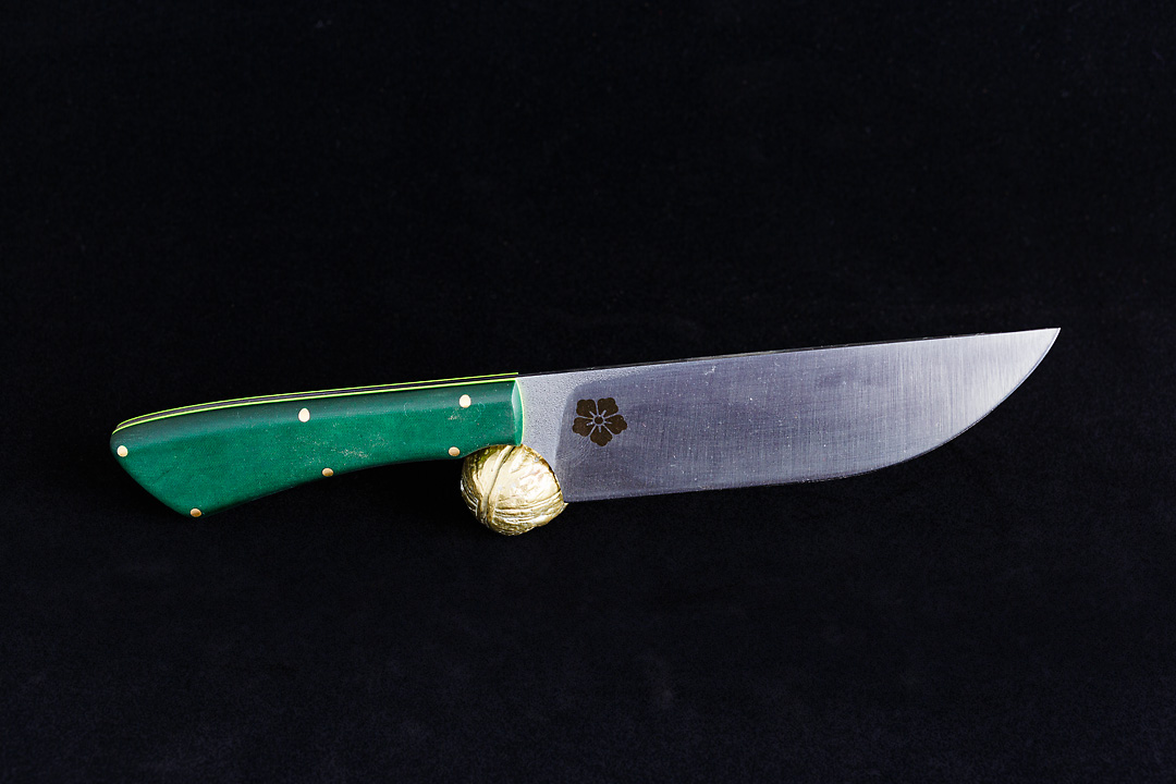 Product photography of a knife with the Kanji Knives logo ingraved on a black background. The knife has a green handle and in the core is a bright green line visible. The knife rests on a golden walnut.