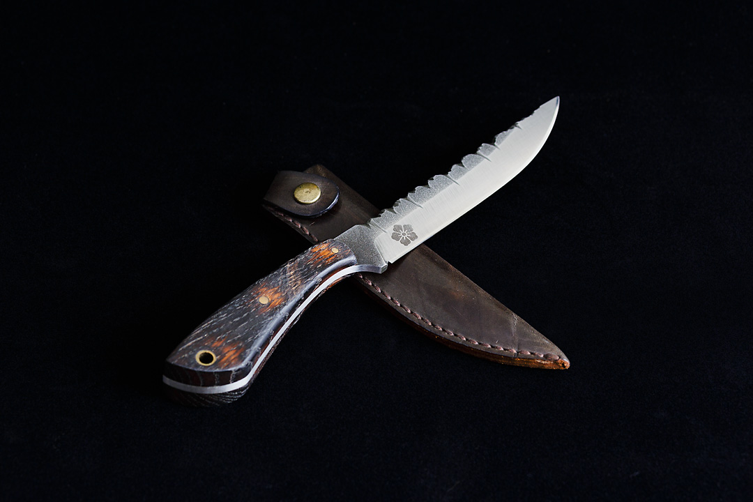 Product photography of a hunting knife with the Kanji Knives logo ingraved on a black background. The knife has a wooden, brown handle. The top of the blade has a irregular relief. The knife rests on a brown leather sheath.