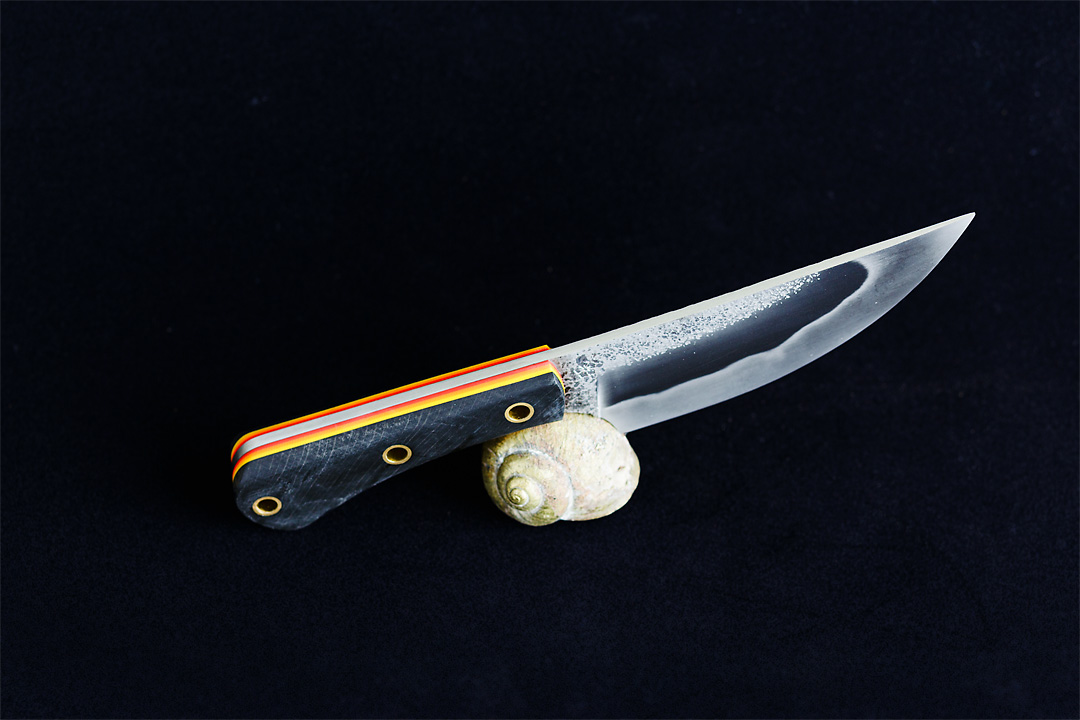 Product photography of a knife with the Kanji Knives logo ingraved on a black background. The knife has a black handle and in the core is a bright yellow line next to a green line visible. The knife rests on a golden shell.