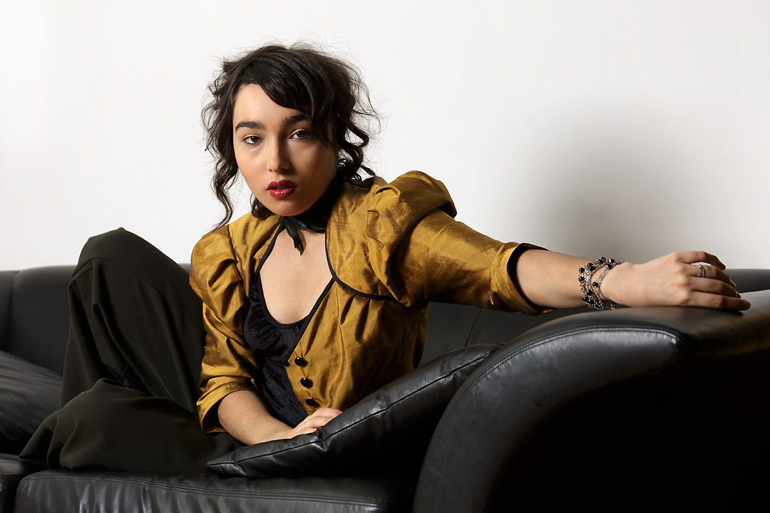 Portrait photography of fashion designer Alex Vanova wearing red lipstick, golden eyeshadow and a golden jacket. She turns and leans towards the camera. Her left arm is streched and her left hand rests on the armrest of a black couch. Around her left wrist is a bracelet with blue gem stones. Her necklace is a choker decorated with feathers.