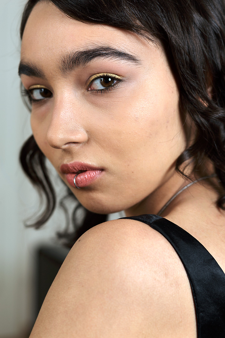 Close-up portrait photography of fashion designer Alex Vanova. She poses with a lip piercing in front of a black cupboard. She wears a shiny oriental necklace and black eye-liner. Her dark brown eyes look directly into the camera.