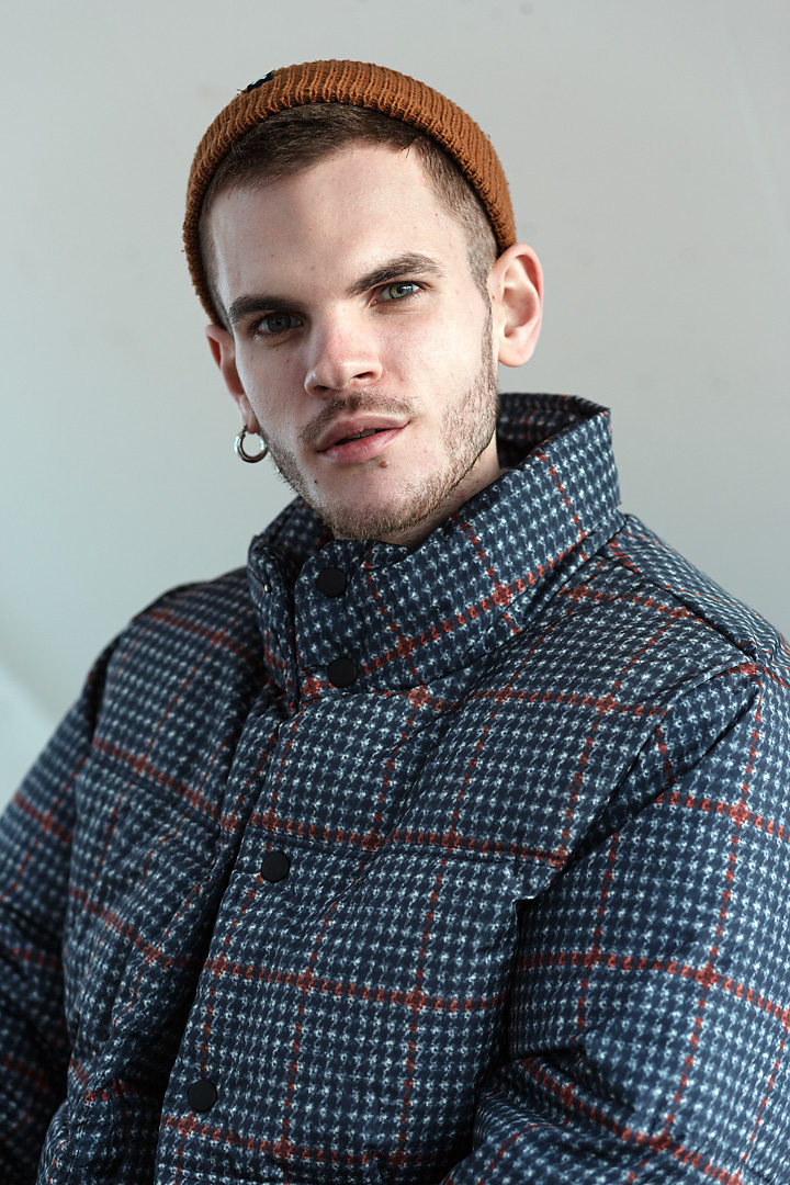 Portrait photography of Brizzi Bremse, a German social media influencer.  His right ear is pierced by a steel earring. His head is slightly tilted to the right. His left side is lid by a very soft light, whereas his right side is a bit darker. He wears a mustard-brown cap and a chequered winter jacket.