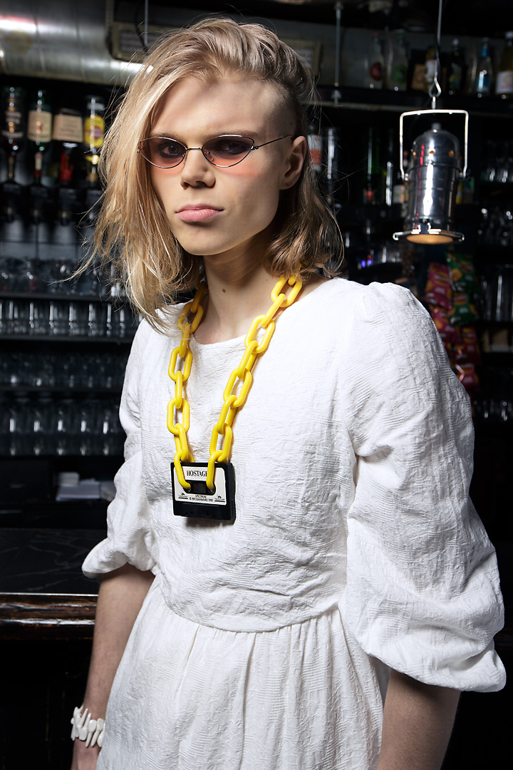 Portrait of Lasse, a young man with long blond hair. He wears a white dress, a yellow plastic-chain necklace with a black tape cassette, a bracelet of white plastic teeth and pink tainted sunglasses. He stands in front of a bar.