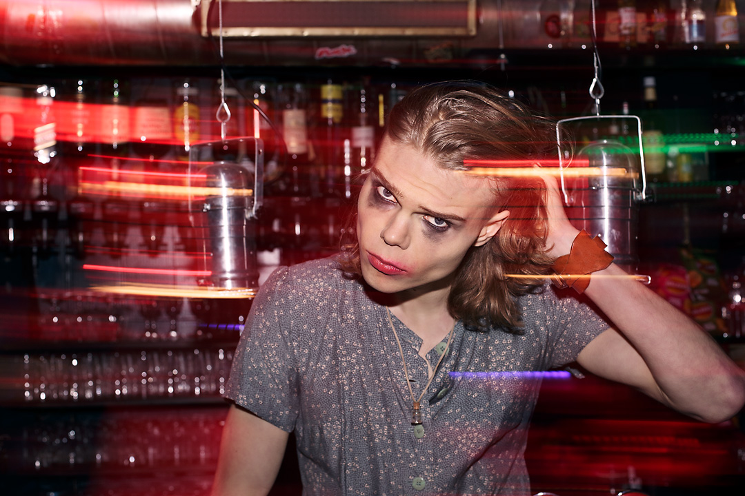 Portrait of Lasse, a young man, who reaches with his left hand to his longer blond hair. He wears a blue-grey-ish dress with a print of small flowers. Both his red lipstick as his black eye shadow are smeared. The background is smeared, but is still recognasible as a bar.