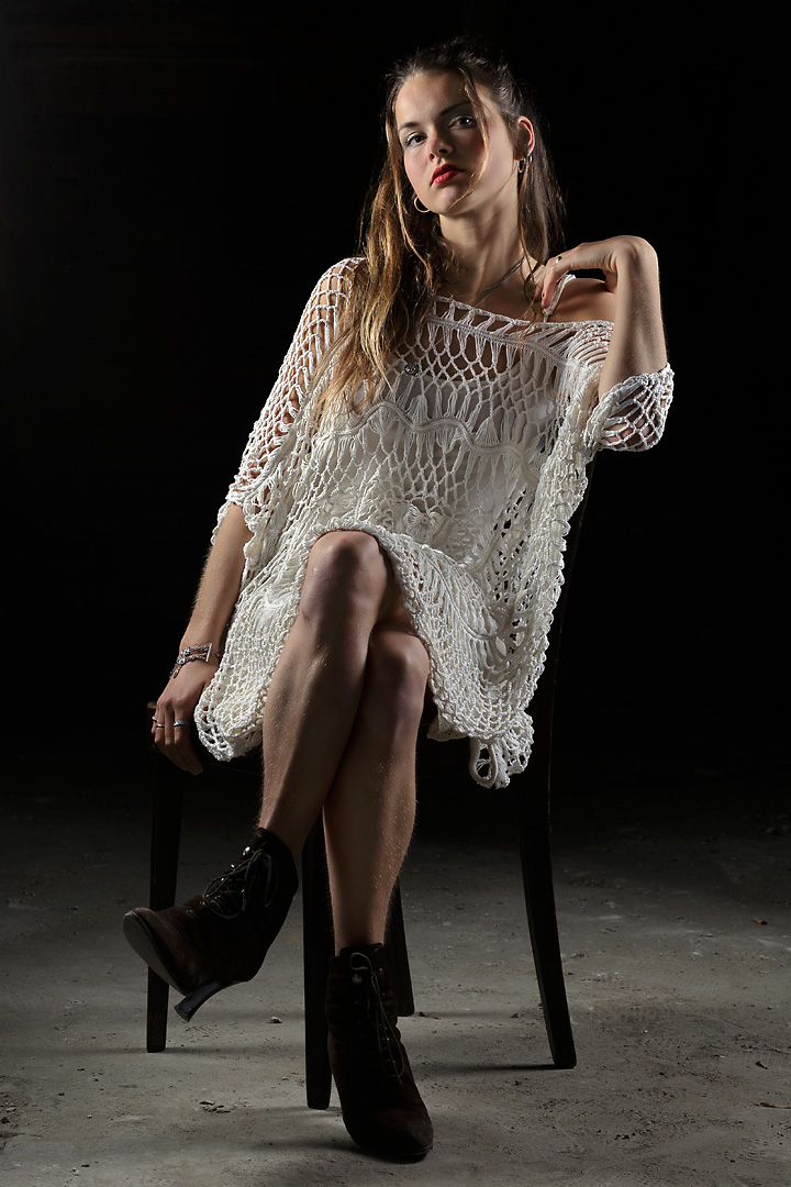 Portrait photography of Lotte, a young woman sitting in front of the camera. She wears a white, knitted dress. With her left hand she touches her left shoulder. Her legs are crossed and she wears small boots.