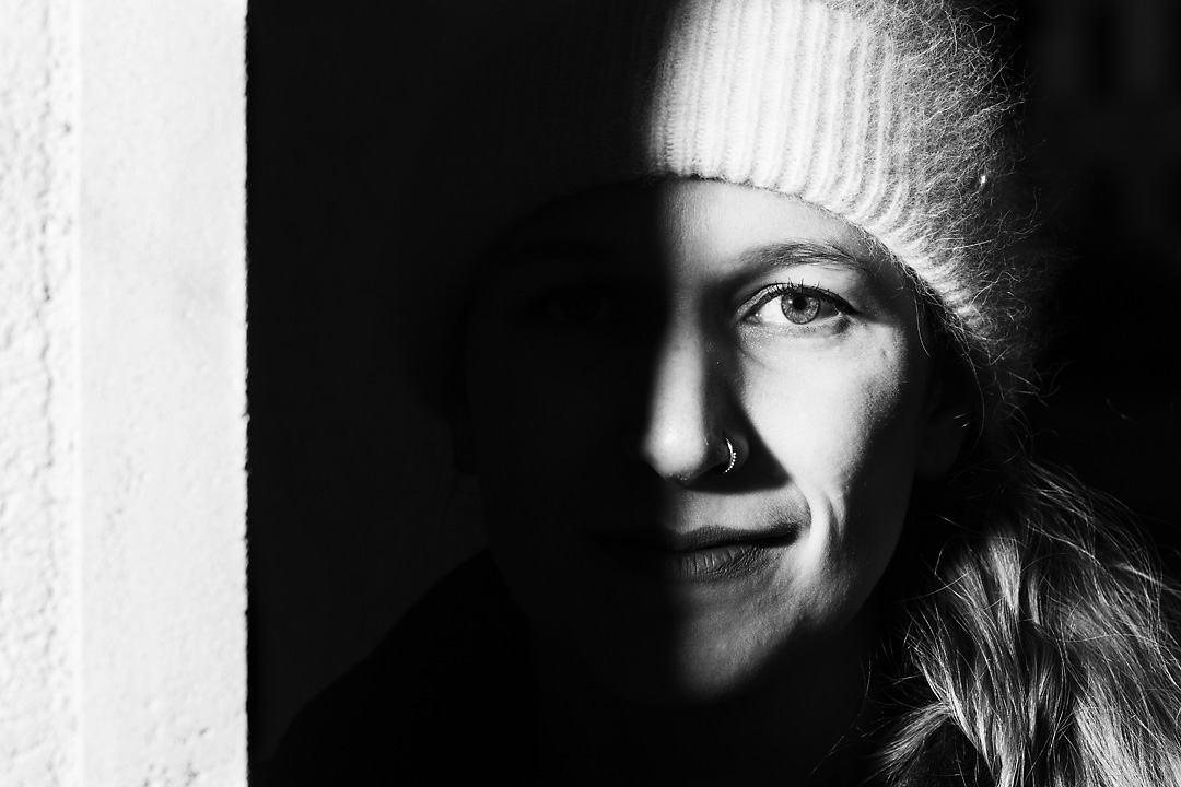 Black and white portrait photography of a woman wearing a woolen cap. Only the right side of her face is visible as the other side is hidden in the shadows of a wall.