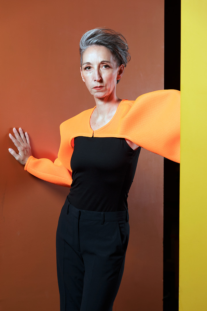 Portrait photography of Romana, a woman with short grey hair in a corridor. The background is orange and the foreground is yellow. Over her black tank top she wears a bright orange top designed by Fred Kobayashi. Her right hand rests against the background, while her left arm is stretched forward towards the opposite wall.