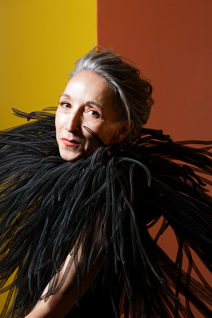 Portrait photography of Romana, a woman with short grey hair in front of a yellow and orange background. She wears a top piece of many small black tubes designed by Fred Kobayashi. She turns her head to the left and looks directly into the camera.