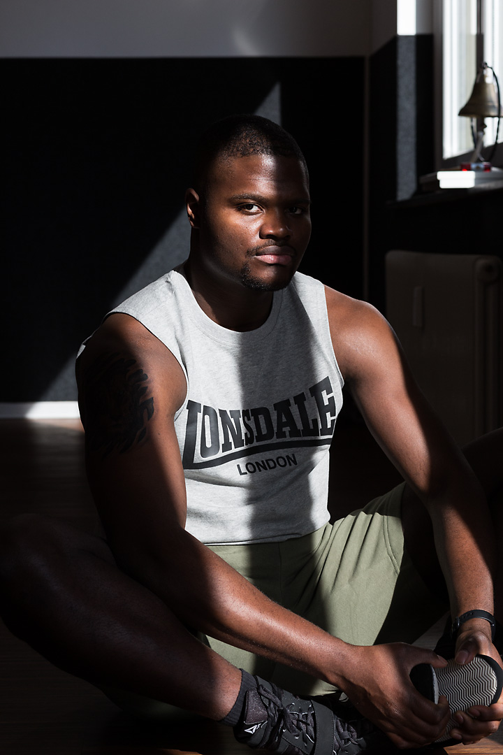 Portrait photography of Oluwaseun Salami, an Austrian professional boxer. He sits on a wooden gym floor and streches his groin. Half of his face is covered by the shadow of a window.