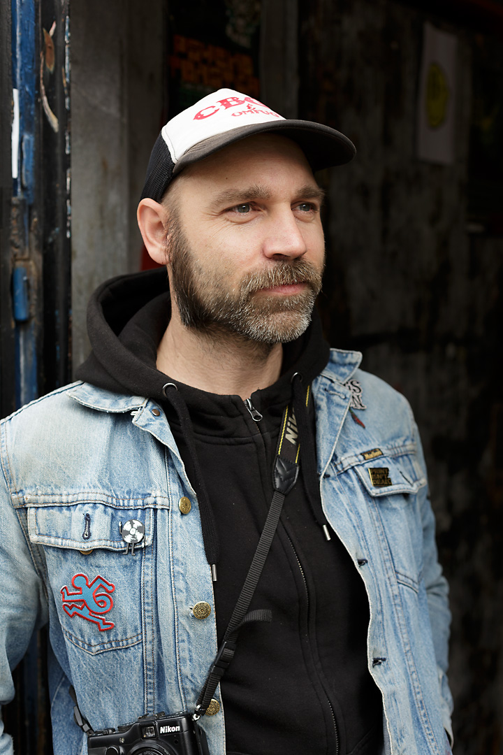 Portrait photography of Zellerluoid, an Austrian screenprinter and illustrator based in Barcelona. He stands in a doorway and looks outside the frame. He wears a jeans jacket, a black hoodie and a black and white trucker cap.