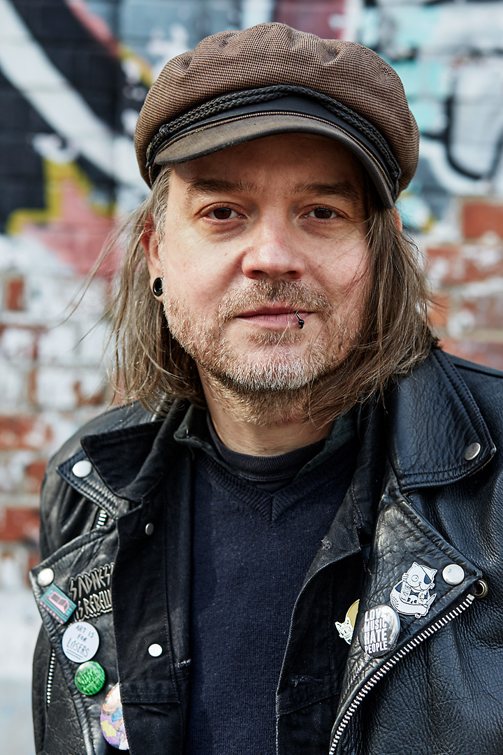 Portrait photography of Stay Gold Grafix, an Austrian illustrator and graphic designer. He wears a black leather jacket with different pins and a brown cap.