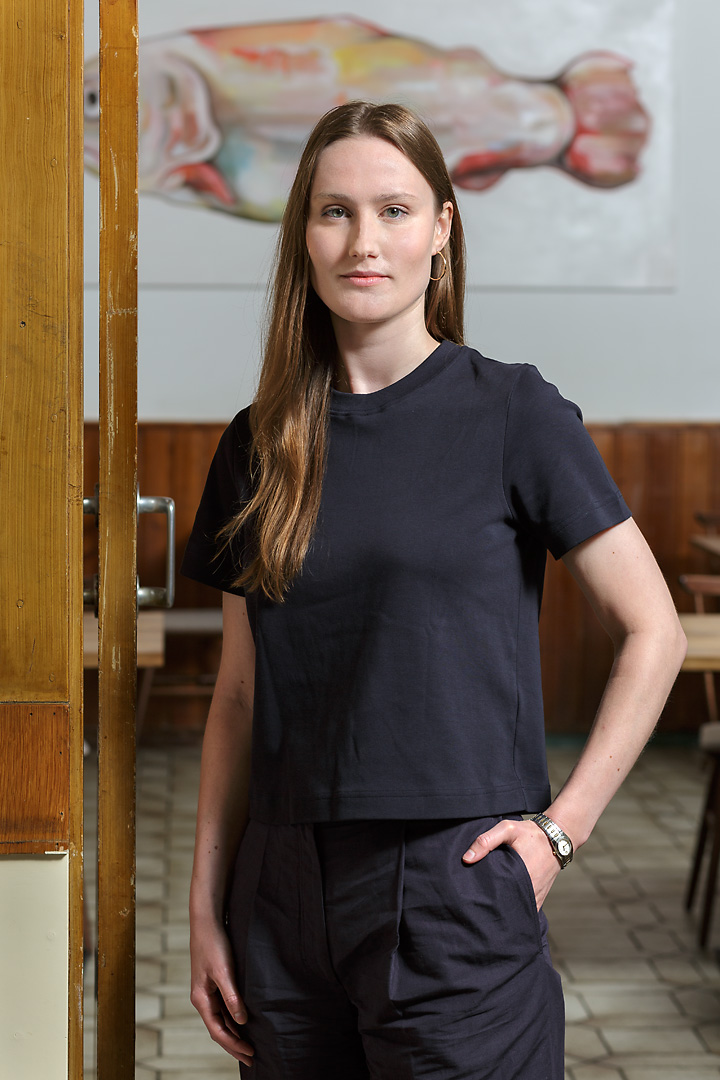 Portrait photography of a waitress at Reznicek. She wears a black t-shirt and black trousers. She stands in a doorframe and in the background is a large painting of a fish. Her long brown hair is rests over her right shoulder.