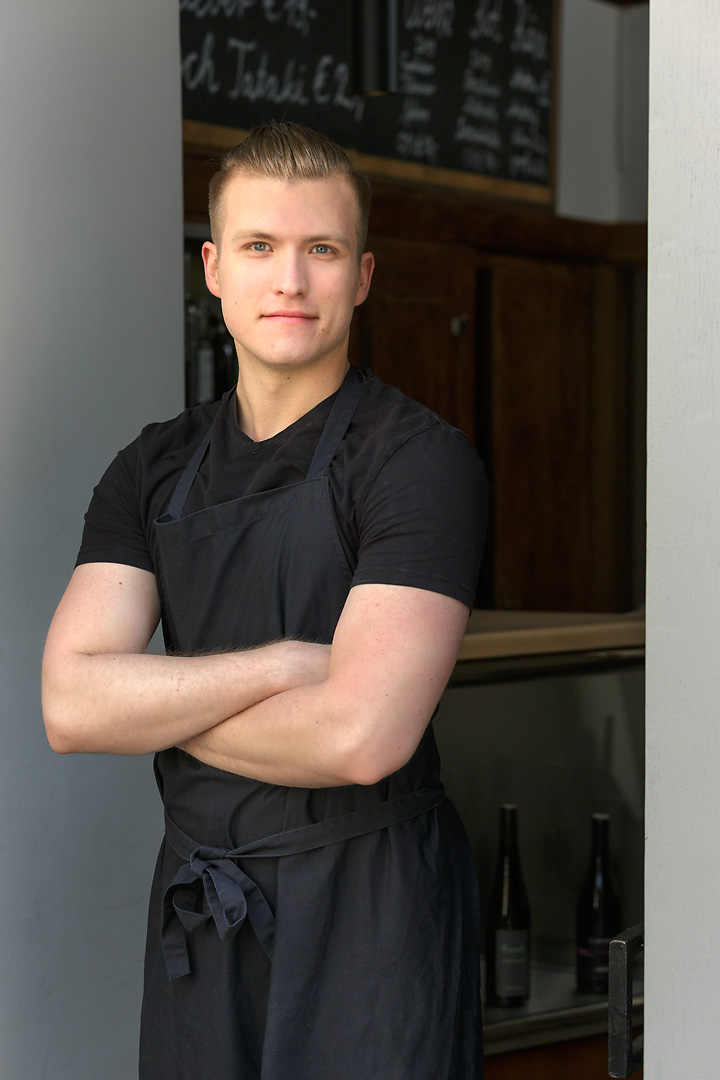 Portrait photography of a cook at Reznicek. He wears a black t-shirt and a black apron. He stands in the doorframe of the entrance of the restaurant. His arms are crossed infront of his chest.