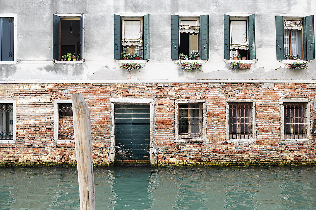 Frontal streetphotography of a facade in Venice, Italy. The picture is divided in three lines. The bottom line is the canal with its green-blueish water and a reflection of the building. The middle line is the ground floor of the building with bared windows, brick facade and a wooden green door. The top line is the first floor of the building with light greyish plaster and wooden green window shutters.