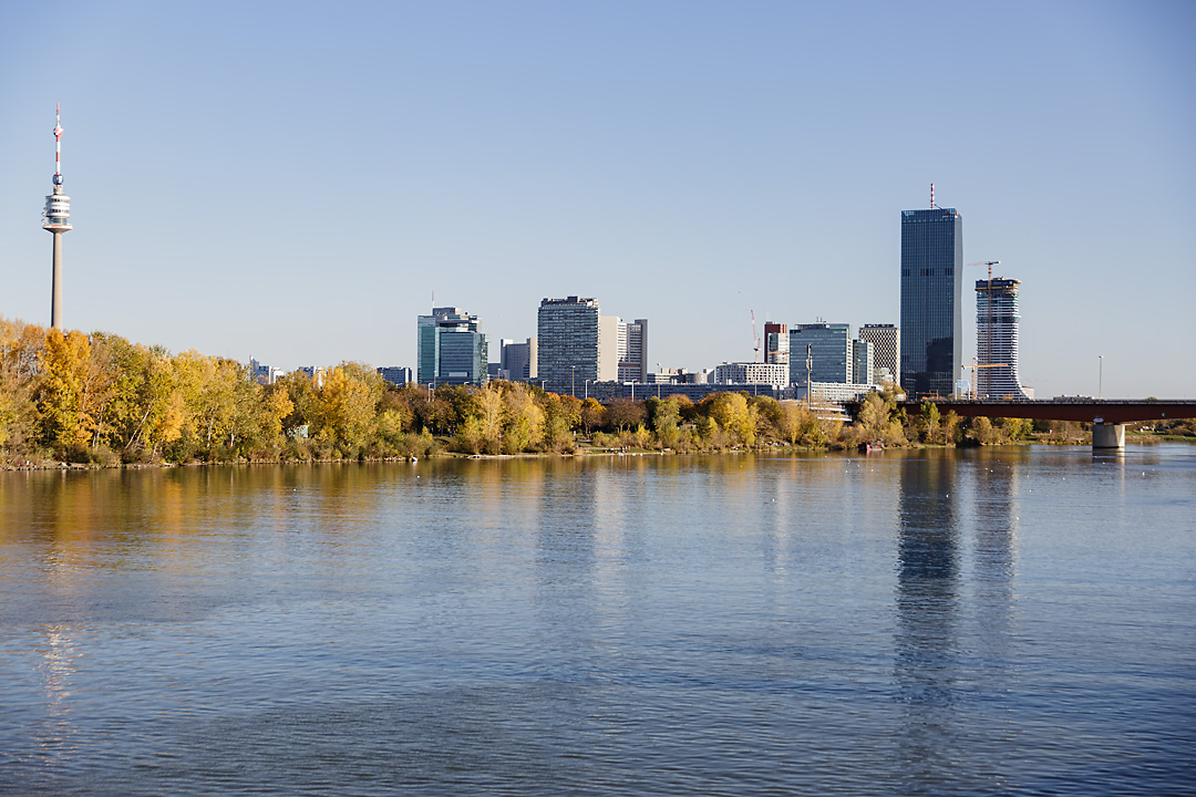 Streetphotography of the Vienna International Center. On the left edge of the picture is the Donauturm. The Danube builts the foreground and a leading line of yellow trees leads to the buildings. 