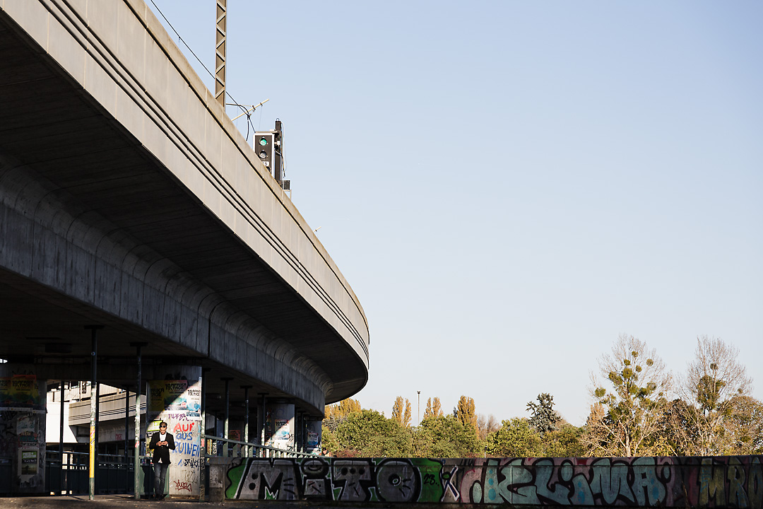 Streetphotography of the U6 metro bridge crossing the Neue Donau. The bridge starts in the top left corner and goes to the center of the picture. Under the bridge is a small pocket of sunlight, where a by-passer looks on a device in his hands.