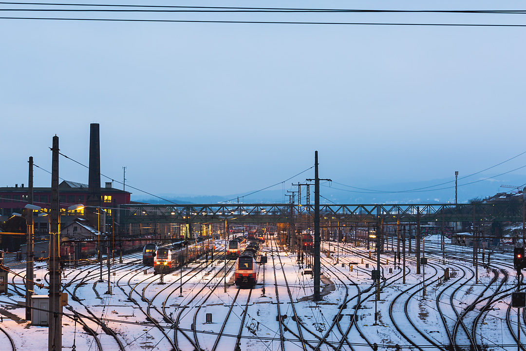 Streetphotography of the Westbahnhof Traktion in winter. From an elevated perspective the picture shows train tracks covered by a bit of snow. In the middle of the picture going from left to right is the Rustensteg Bruecke.