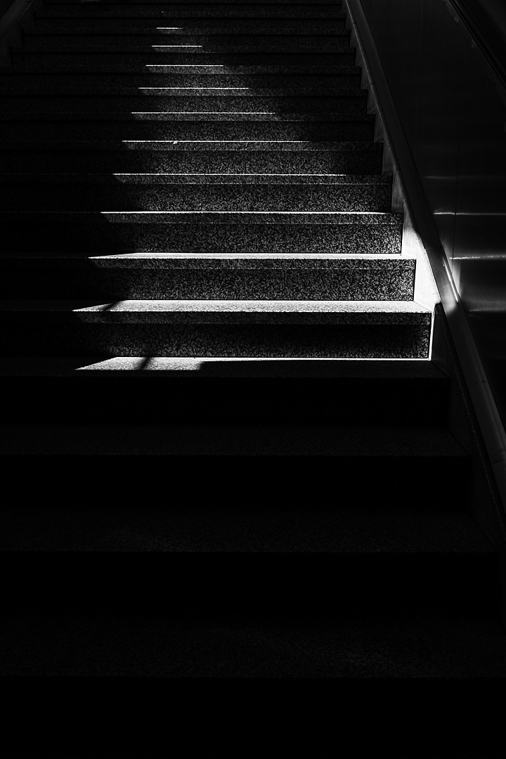 Black and white streetphotography of a staircase. The picture is very dark, except for a triangle, which is hit by bright sunlight.