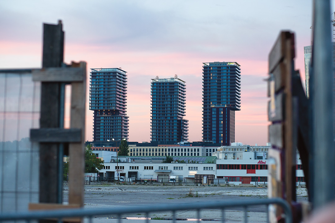 Streetphotography from the Triiiple buildings in Vienna. This picture is taken from an alternatively-used plot in St. Marx. The buildings are framed by an DIY-wooden fence entrance and against the backdrop of a pink evening sky.