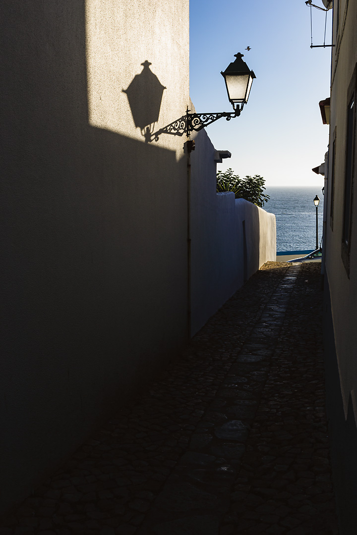 Streetphotography of the Ericeira, Portugal. A street lantern in a small alley casts its shadow late afternoon shadow on a wall. In the background the atlantic ocean is visible.