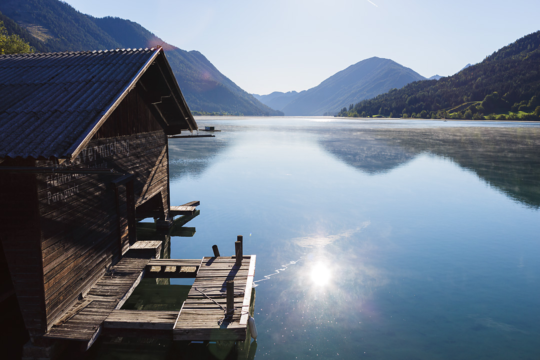 Streetphotography of a sunset on Weissensee in September. The water reflects the sun and the mountains in the horizon. On the left side is a wooden cabin.