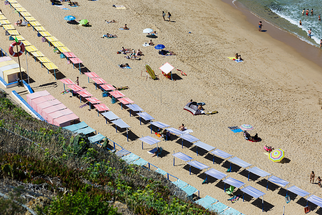 Streetphotography of Praia do Sul in Ericeira, Portugal. In the top right corner some people a standing in the ocean. From the top left corner to the bottom right corner are diagonal lines of colourful sun covers. The foreground in the left bottom corner shows the edge of a cliff covered with vegetation.