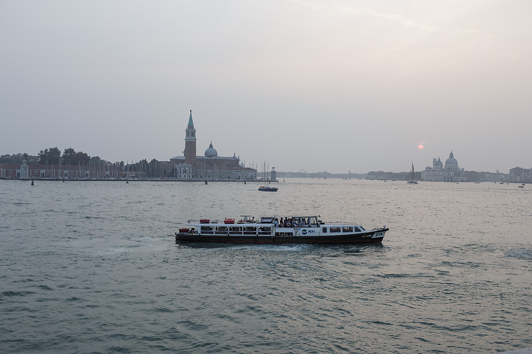 Streetphotography of Venice with a hazy sunset in the right back. In the foreground is a boat and in the background the Basilica di San Marco and the Abbazia di San Giorgio Maggiore.