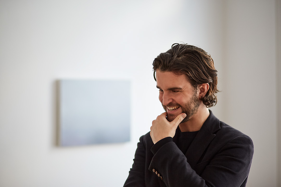 Portrait photography of the painter Tom Van Puyvelde next to his Brume IV in the Common Sense Art Gallery in Vienna. The artists is positioned on the right side. His left hand touches his chin as he smiles and looks outside of the frame.