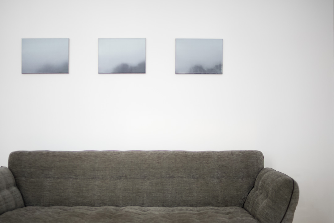 A static photography of three paintings by Tom Van Puyvelde in the Common Sense Art Gallery in Vienna. The paintings Brume V, Brume VI and Brume VII are in line and below these paintings is a green couch, which fills almost the bottom third of the picture.