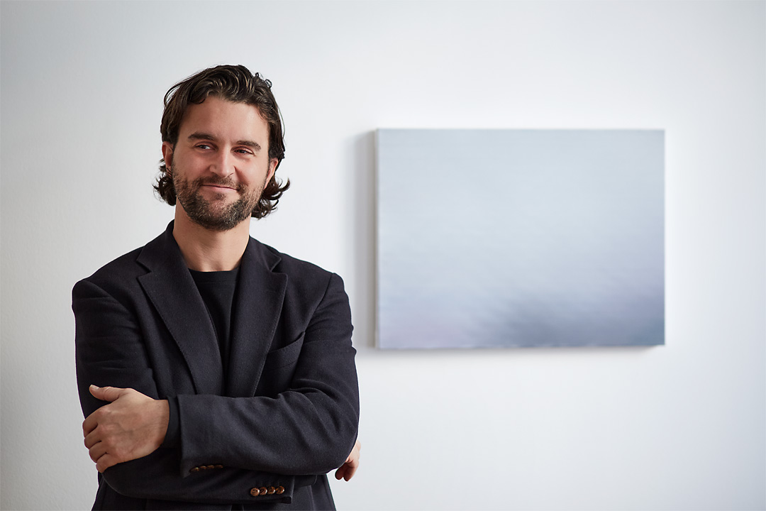 Portrait photography of the painter Tom Van Puyvelde in front of his Brume IV in the Common Sense Art Gallery in Vienna. The artists is positioned in the left and has his arms crossed. He looks outside the frame and a smile forms on his face.